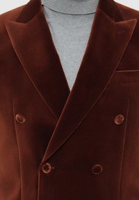 Dvigubo užsegimo rudos veliūro švarkas uždėtas ant šviesiai pilkos megztinio su apvaliu kaklu, matomi atvartai ir veliūro uždengti mygtukai.