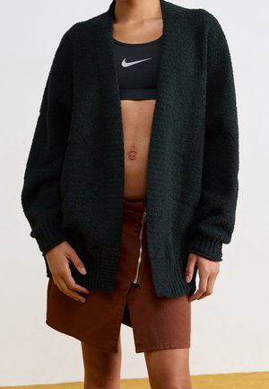 Personne portant un cardigan noir duveteux sur un soutien-gorge de sport noir Nike et une jupe marron avec une fermeture éclair à l'avant, debout devant un fond uni.