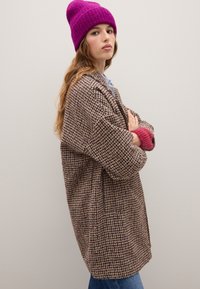 Abrigo grande de color marrón, púrpura y crema con un estampado, con dos bolsillos, superpuesto a una camisa azul clara y combinado con un gorro de punto rosa.
