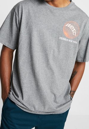 Homme portant un t-shirt gris à manches courtes avec un graphique sphérique orange et le texte "Wesc Stockholm / New York", mains dans les poches d'un pantalon bleu foncé.
