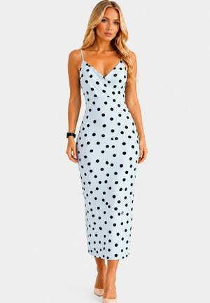 Femme portant une robe bleu clair sans manches longue jusqu'aux chevilles avec des pois noirs, associée à des talons nude et une montre-bracelet noire.