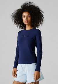 Mlada ženska z kodrastimi lasmi, oblečena v temno modro majico z dolgimi rokavi z napisom "Red Bull Racing" in svetle denim kratke hlače, na sivem ozadju.