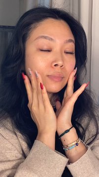 Frau mit mittellangen schwarzen Haaren und ohne Make-up, die einen beigen Pullover trägt und ihr Gesicht mit rot lackierten und gestreiften Fingernägeln sowie Armbändern berührt.