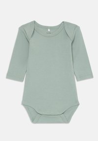 Långärmad babymössa i mjuk salvia grön bomull. Har en rundad halsringning och tryckknappar i botten för enkel påklädning.