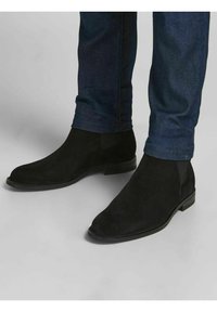 Czarne zamszowe botki za kostkę o gładkim wykończeniu, z zaokrąglonym noskiem i niskim, warstwowym obcasem, zestawione z ciemnymi dżinsowymi spodniami.