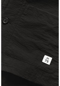 Black shirt with a textured fabric, featuring a button closure, a patch pocket, and a white label displaying "M 73."