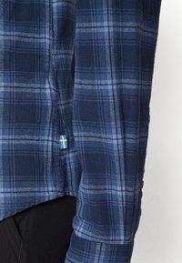 Cămașă din flanel în carouri albastru marin cu dungi albastre deschise, având un etichetă cu steagul suedez albastru și galben aproape de tiv. Textură moale, croi relaxat.