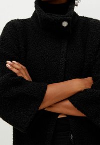 Cappotto nero, soffice e di taglia oversize con un colletto alto e una chiusura a bottone lucido singolo. Presenta maniche ampie e un tessuto testurizzato.