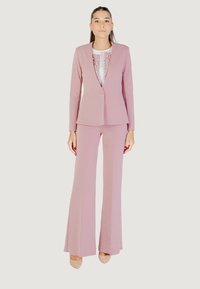Donna in piedi con indosso un blazer su misura color malva e pantaloni larghi abbinati, con tacchi a punta beige, su uno sfondo neutro.