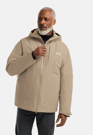 Mann mit grauem Bart trägt eine beige Kapuzenjacke mit Jack Wolfskin-Logo und zieht den Reißverschluss vor einem einfarbigen weißen Hintergrund hoch.