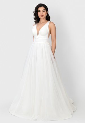 Vrouw die een mouwloze witte jurk draagt met een diepe V-hals en een volle tule rok, staand tegen een effen lichte achtergrond.