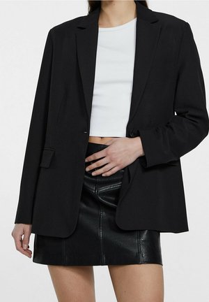 Person trägt einen schwarzen Blazer über einem weißen Cropped-Top und einem schwarzen Leder-Minirock, mit einer Hand auf der Taille.