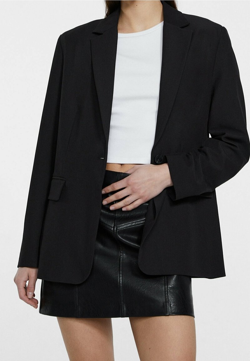 Personne portant un blazer noir sur un top court blanc et une mini-jupe en cuir noire, une main posée sur la taille.