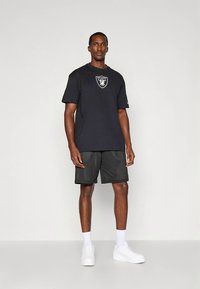 Svart t-shirt i bomull med Raiders-logotyp på bröstet, kombinerad med svarta meshshorts. Vita sneakers och vita strumpor i crew-längd fulländar stilen.