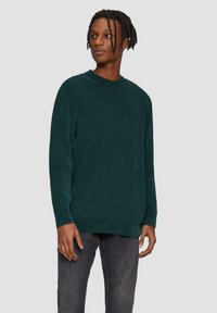 Dunkelgrüner gerippter Pullover mit Rundhalsausschnitt, langen Ärmeln und lockerer Passform, kombiniert mit dunkelgrauen Jeans. Weiche Textur, minimalistisches Design.