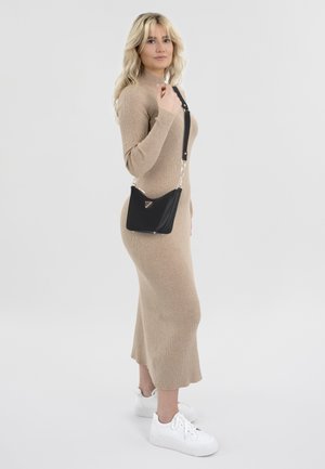Zwarte schoudertas met een driehoekig logo, zilveren kettingriem; model in een beige geribd langmouwkleed en witte sneakers.