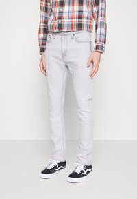 Muž v lehce šedých slim-fit džínách, černo-bílých teniskách a dlouhém kostkovaném tričku s červenou, černou a bílou barvou.