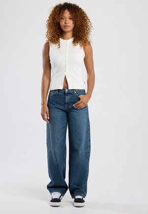 Top blanco sin mangas con cierre frontal de botones, combinado con pantalones vaqueros de mezclilla oscuros y holgados. Calzado: zapatillas negras y blancas.