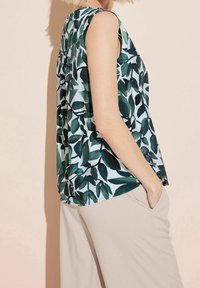 Sleeveless top with green and black leaf pattern worn with light beige pants, hand in pocket, shown from side on neutral background.