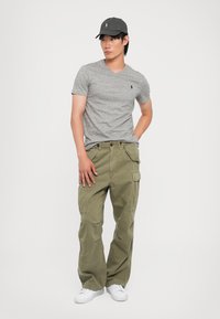 Γκρι t-shirt με v λαιμόκοψη και μικρό λογότυπο, ελαιόχρωμες cargo παντελόνες με πολλές τσέπες και ένα σκούρο καπέλο. Λευκά αθλητικά παπούτσια ολοκληρώνουν το σύνολο.