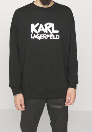 Mężczyzna w czarnej bluzie z białym napisem "KARL LAGERFELD" na piersi, w zestawie z czarnymi spodniami na jednolitym tle.