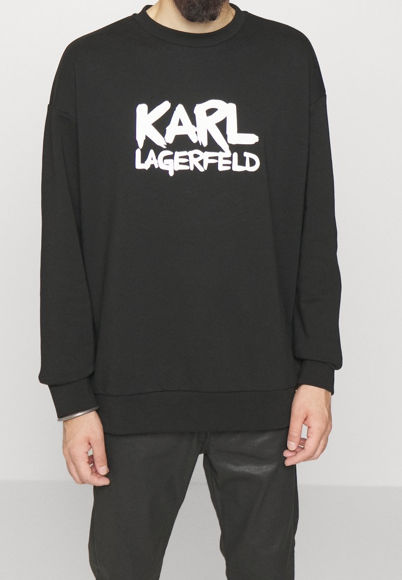 Homme portant un sweat-shirt noir avec le texte blanc « KARL LAGERFELD » sur la poitrine, associé à un pantalon noir sur un fond uni.