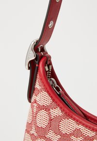 Borsa a spalla in pelle rossa con motivo tessile intrecciato, caratterizzata da hardware argentato e una forma curva. Dettaglio con zip e accenti a contrasto sulla tracolla.