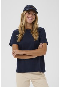 Nasmejana ženska z dolgimi blond lasmi, oblečena v temno modro majico in kapo, bež hlače, stoji s prekrižanimi rokami pred belo ozadje.