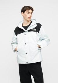 Młody mężczyzna w białej i czarnej kurtce The North Face z rękami w kieszeniach, patrzący na bok na jednolitym tle.