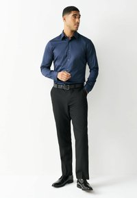 Man staat met hand in zijn zak, draagt een donkerblauw overhemd met knopen, zwarte nette broek, zwarte riem en zwarte nette schoenen, kijkend naar de zijkant.