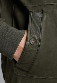 Chaqueta de cuero verde oscuro con puños de punto acanalado, que presenta un bolsillo con un acento de botón metálico redondo y costuras detalladas.