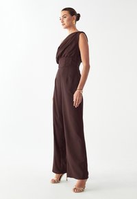 Rjav kombinezon z zavesnim, asimetričnim izrezom, prilagojenim pasom in hlačnicami širokih nog. Znahuje se po gladki tkanini in minimalističnem dizajnu.