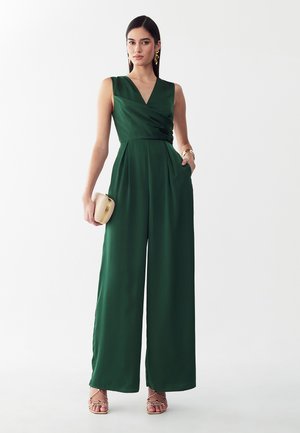 Donna che indossa una tuta verde scuro senza maniche a gamba larga con scollo a V, tiene una pochette beige e indossa orecchini d'oro e sandali.