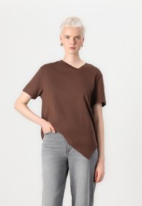 Brun V-hals T-skjorte med korte ermer, med en asymmetrisk hem og myk tekstur. Kombinert med lysegrå bukser.