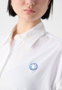 Hvit skjorte med knapper som har et brodert blått smilefjes-logo på venstre bryst. Frisk stoff med klassisk kragedesign.