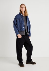 Jeansjacka i mörkblått, med knappstängning, två bröstfickor och avslappnad passform. Parat med grå skjorta och svarta lösa byxor. Svarta skor.