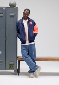 Marineblauw en koraal Nike bomberjack, wit shirt, losse blauwe spijkerbroek, grijze sneakers. Grijze locker op de achtergrond, houten bankje, basketbal erop.