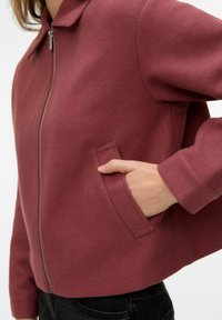 Personne portant une veste zippée bordeaux avec col, main dans la poche latérale, associée à un pantalon noir.