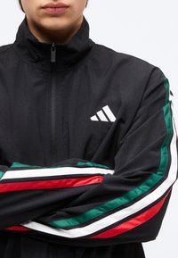 Άτομο που φοράει μαύρο μπουφάν Adidas με φερμουάρ στο κολάρο και κόκκινες, λευκές, πράσινες ρίγες στα μανίκια, με τα χέρια σταυρωμένα.