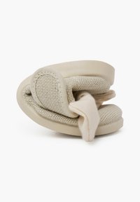 Calzado slip-on beige que presenta un upper de tela texturizada y una suela suave y flexible. El diseño incluye un detalle de nudo en la parte frontal.
