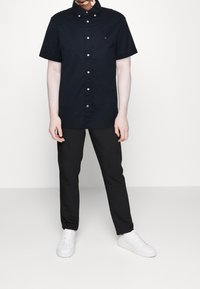 Camicia blu navy a maniche corte con bottoni, in tessuto di cotone, bottoni bianchi, un piccolo logo sul petto, abbinata a pantaloni neri e sneakers bianche.