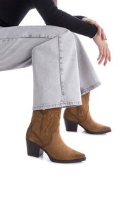 Botas de cowboy de ante marrón con punta afilada, costuras decorativas y un tacón apilado, combinadas con unos vaqueros de pierna ancha grises.