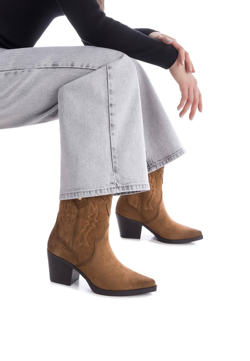 Botas de cowboy de ante marrón con punta afilada, costuras decorativas y un tacón apilado, combinadas con unos vaqueros de pierna ancha grises.