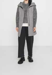 Cappotto grigio senza maniche con cappuccio, morbido maglione grigio a maglia, pantaloni neri larghi con orli sfilacciati e stivali neri con plateau.