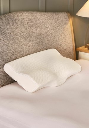 Weißes ergonomisches Kissen mit strukturiertem Bezug auf einem Bett mit braun gepolstertem Kopfteil und einer Nachttischlampe.