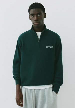 Jeune homme portant un pull vert foncé à fermeture éclair trois-quarts avec un texte blanc et un pantalon gris clair, debout devant un fond blanc uni.