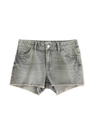 Smilende pige iført en hvid strikket cardigan over en sort top, grå denimshorts og sorte flipflops på en ensfarvet hvid baggrund.