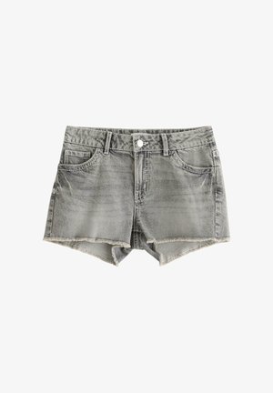 Smilende pige iført en hvid strikket cardigan over en sort top, grå denimshorts og sorte flipflops på en ensfarvet hvid baggrund.