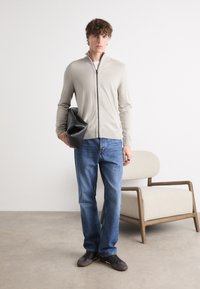 Beige zip-up genser med ribbestrikkede mansjetter, sammen med blå jeans. Modellen holder en svart veske, satt mot en lys bakgrunn.