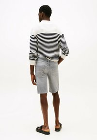 Gestreepte gebreide trui in wit en marineblauw, gedragen met lichtgrijze denim shorts. Model staat met de rug naar de camera, waardoor de platte achterzakdetails zichtbaar zijn. Zwarte sandalen.